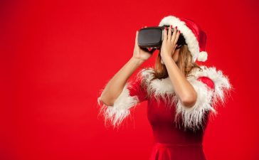 young brunette in santa helper hat wearing virtual reality headset over red background