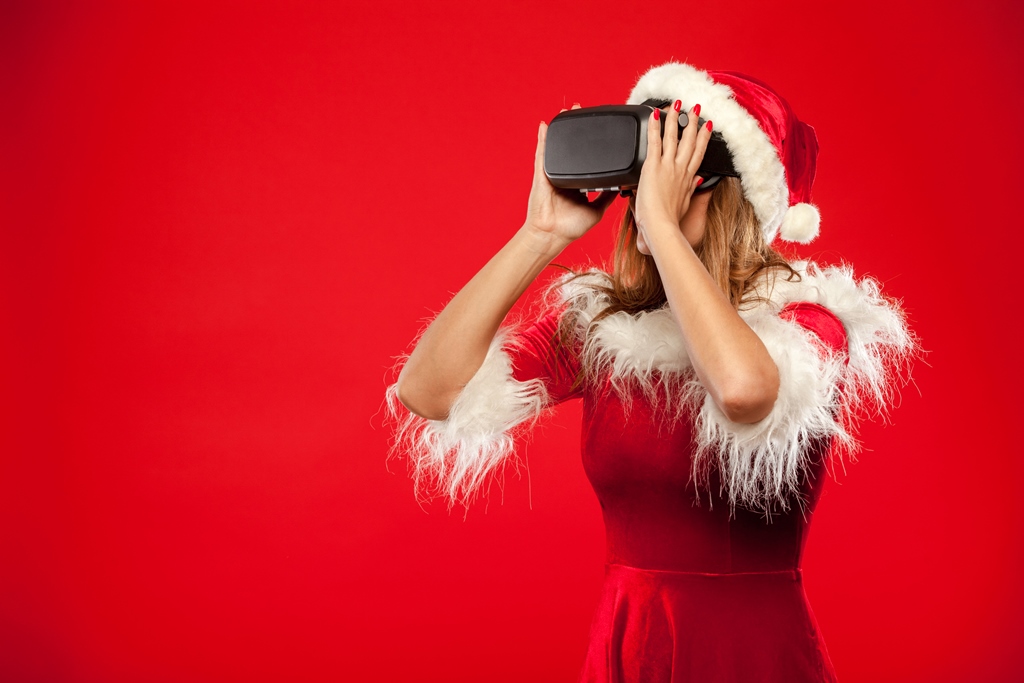 young brunette in santa helper hat wearing virtual reality headset over red background